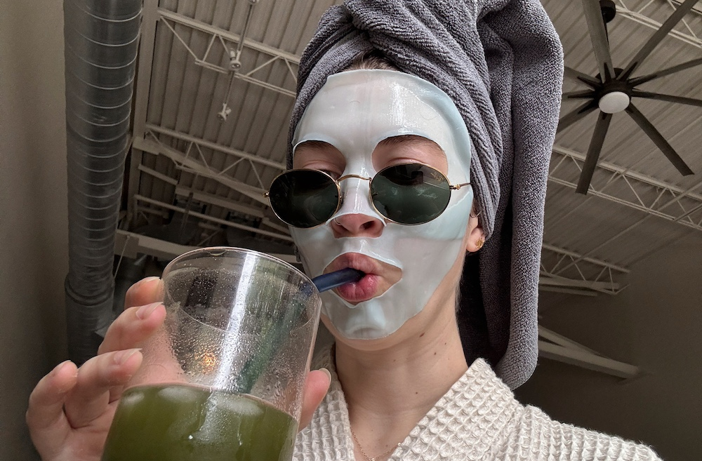 Woman in robe drinks green juice while wearing face mask