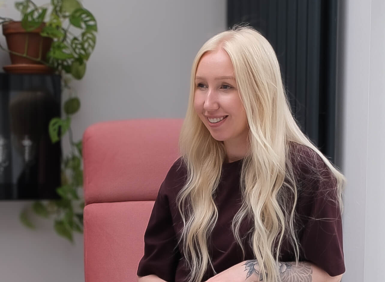 Smiling woman with long blonde hair sits on a pink chair, wearing a dark shirt.
