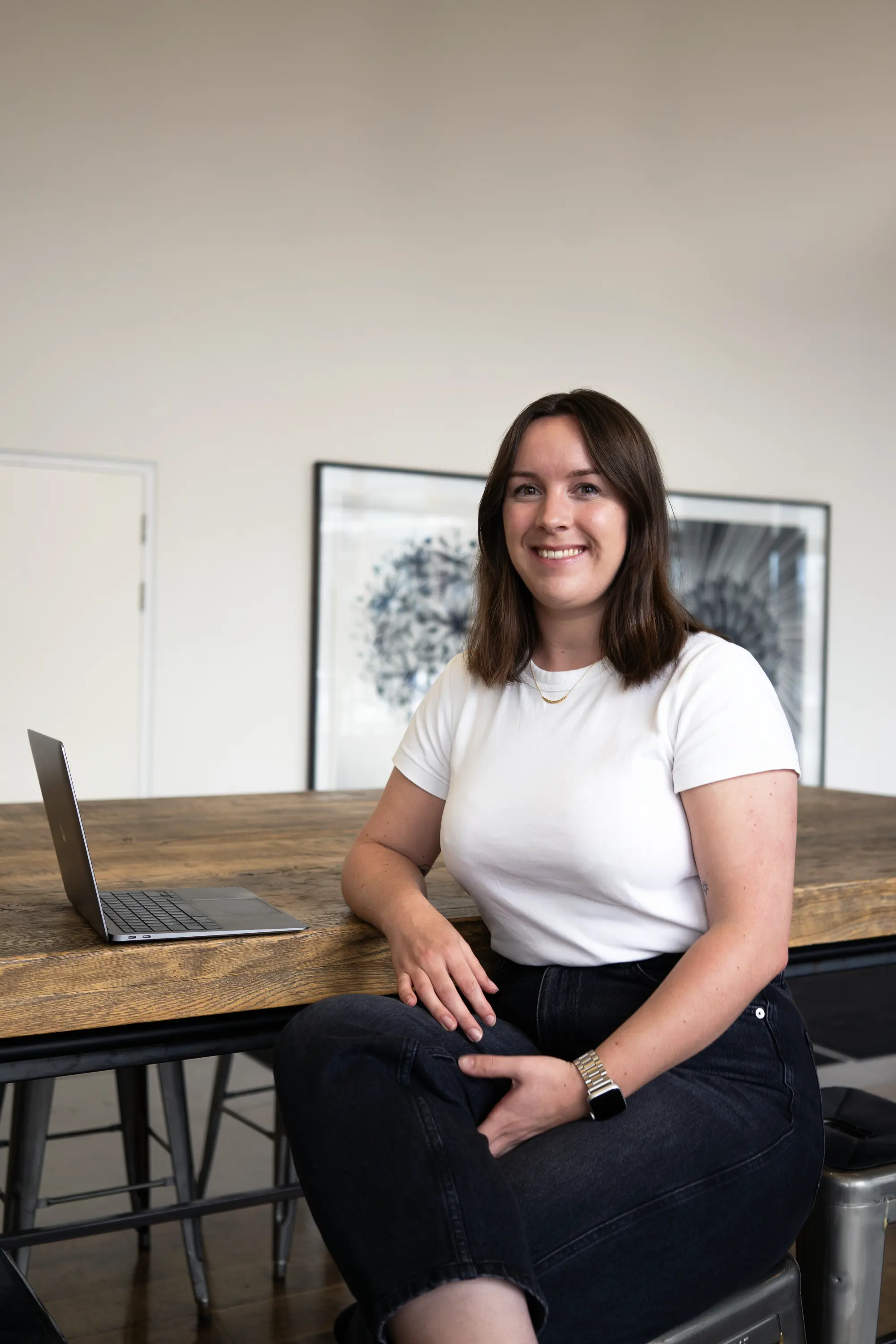 A woman in a white top and black trousers sits at a wooden table with a laptop, smiling.