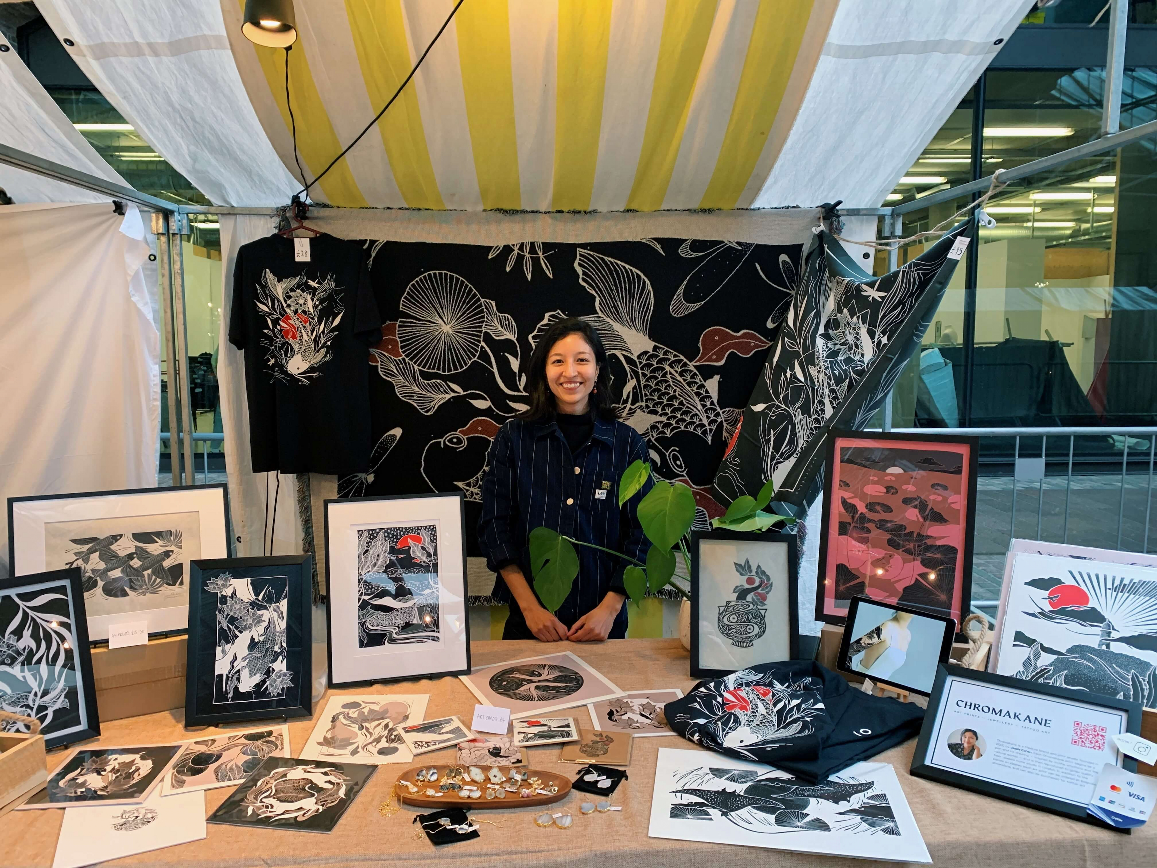A person stands behind a market stall displaying art prints, shirts and small items, with a black and white nature-themed backdrop.