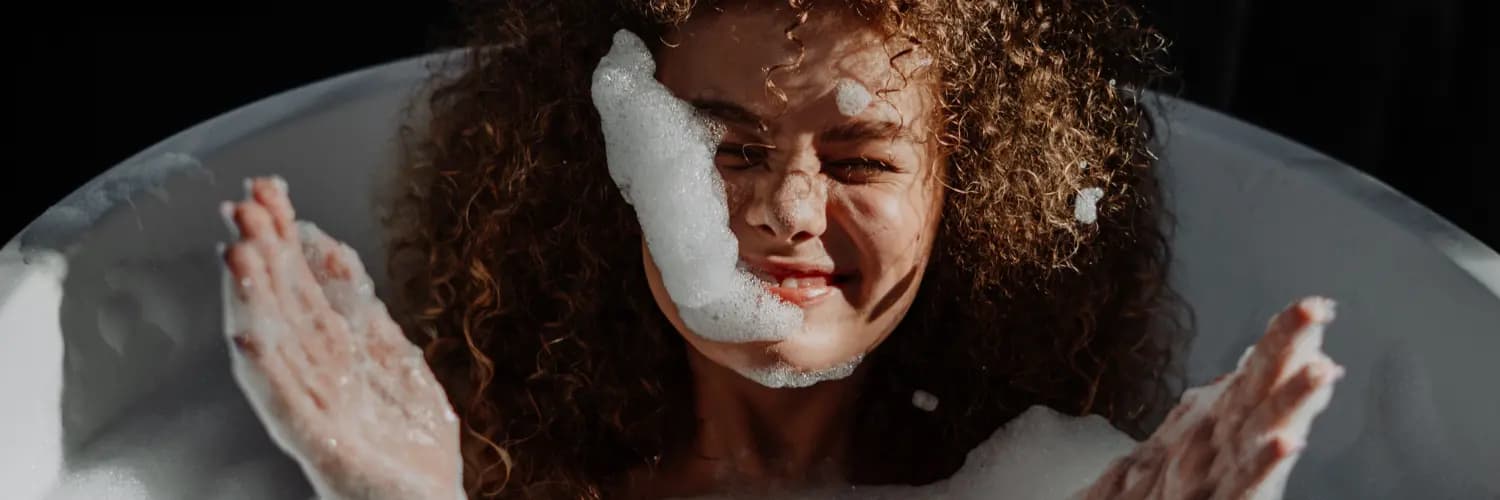 Woman with curly hair laughing and throwing bubbles in the bath