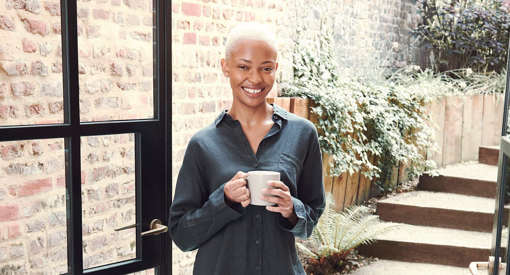 Woman wearing pyjamas stands by open patio doors holding a cup of tea