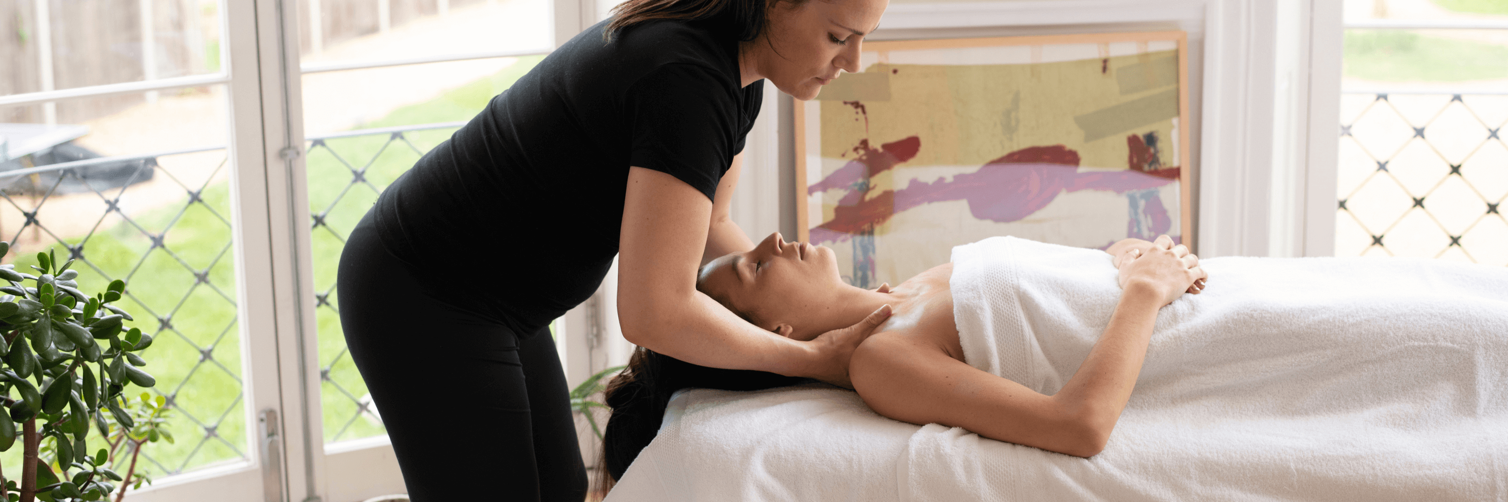 Client lying on massage table on her back draped in white fluffy towel as therapist works on her shoulders