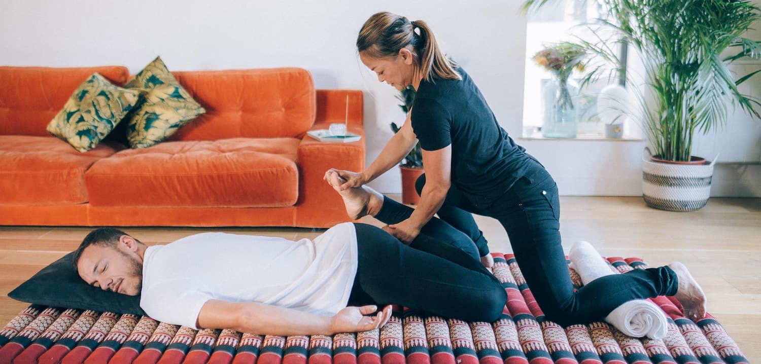 Thai massage therapist stretches client's legs on traditional thai massage mat in London flat