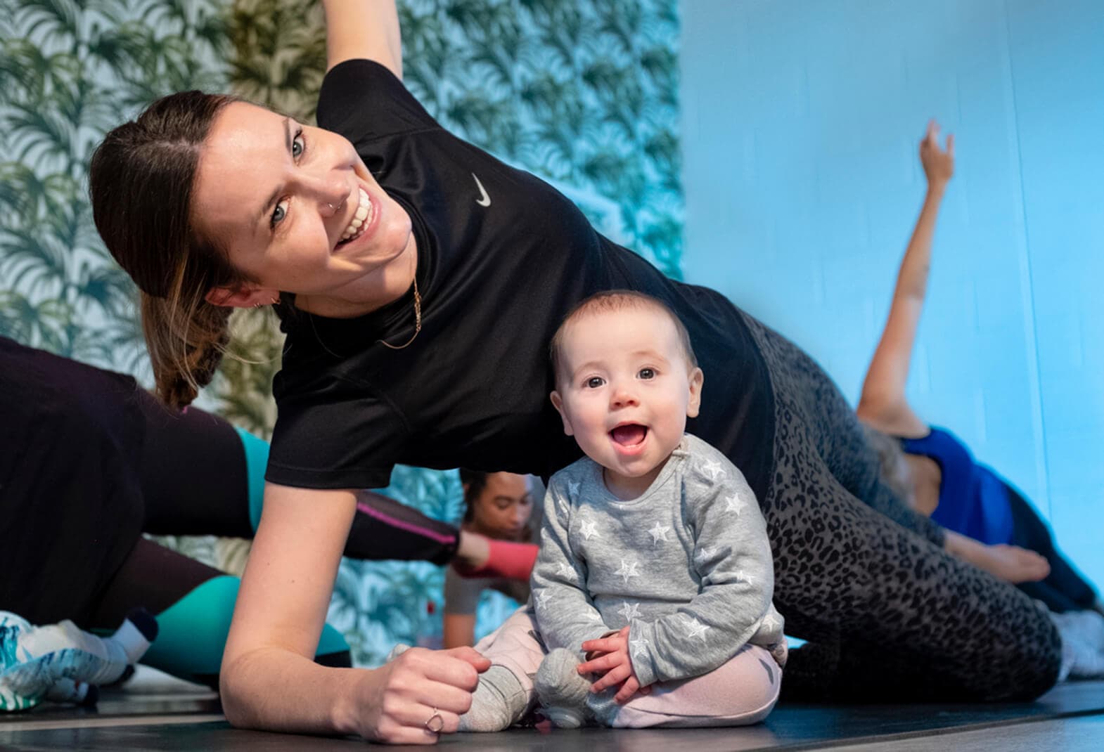 A mum works out on a yoga mat in a gym gear with her baby, both are smiling at the camera 