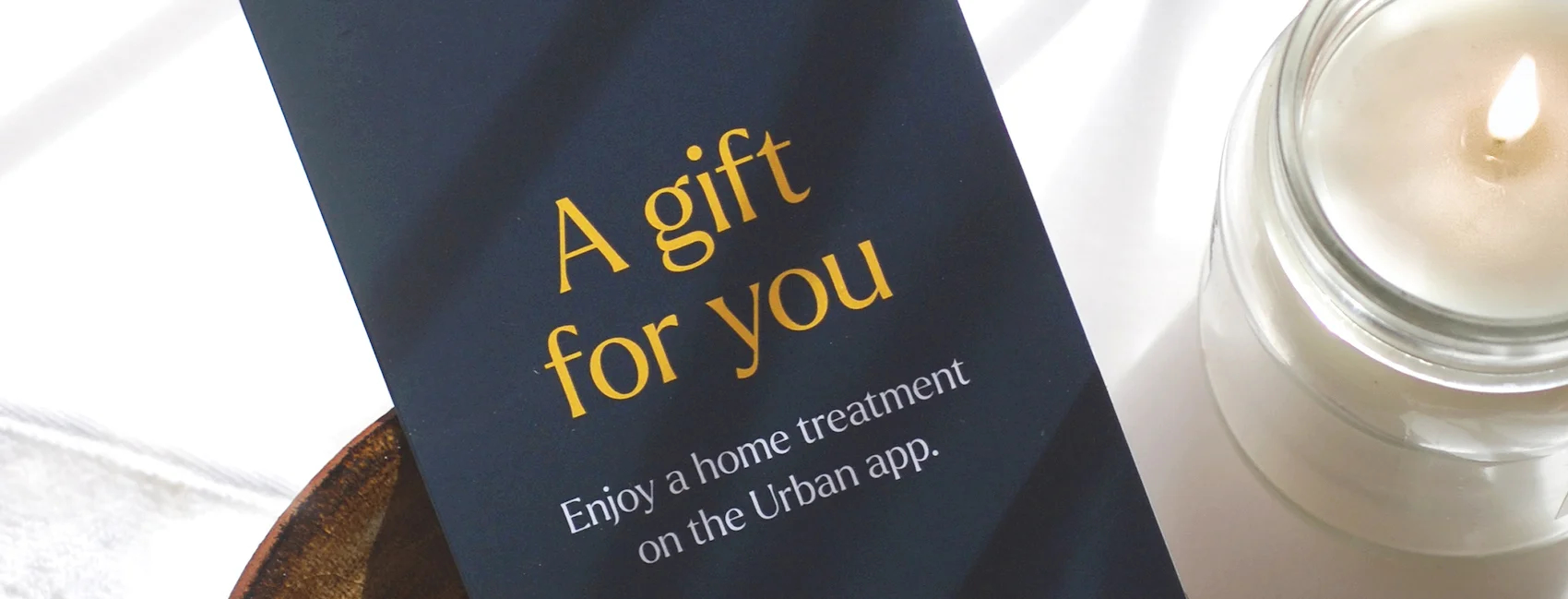 A navy blue massage gift card next to a candle, "gift card reads a gift for you, enjoy a home treatment on the Urban app"