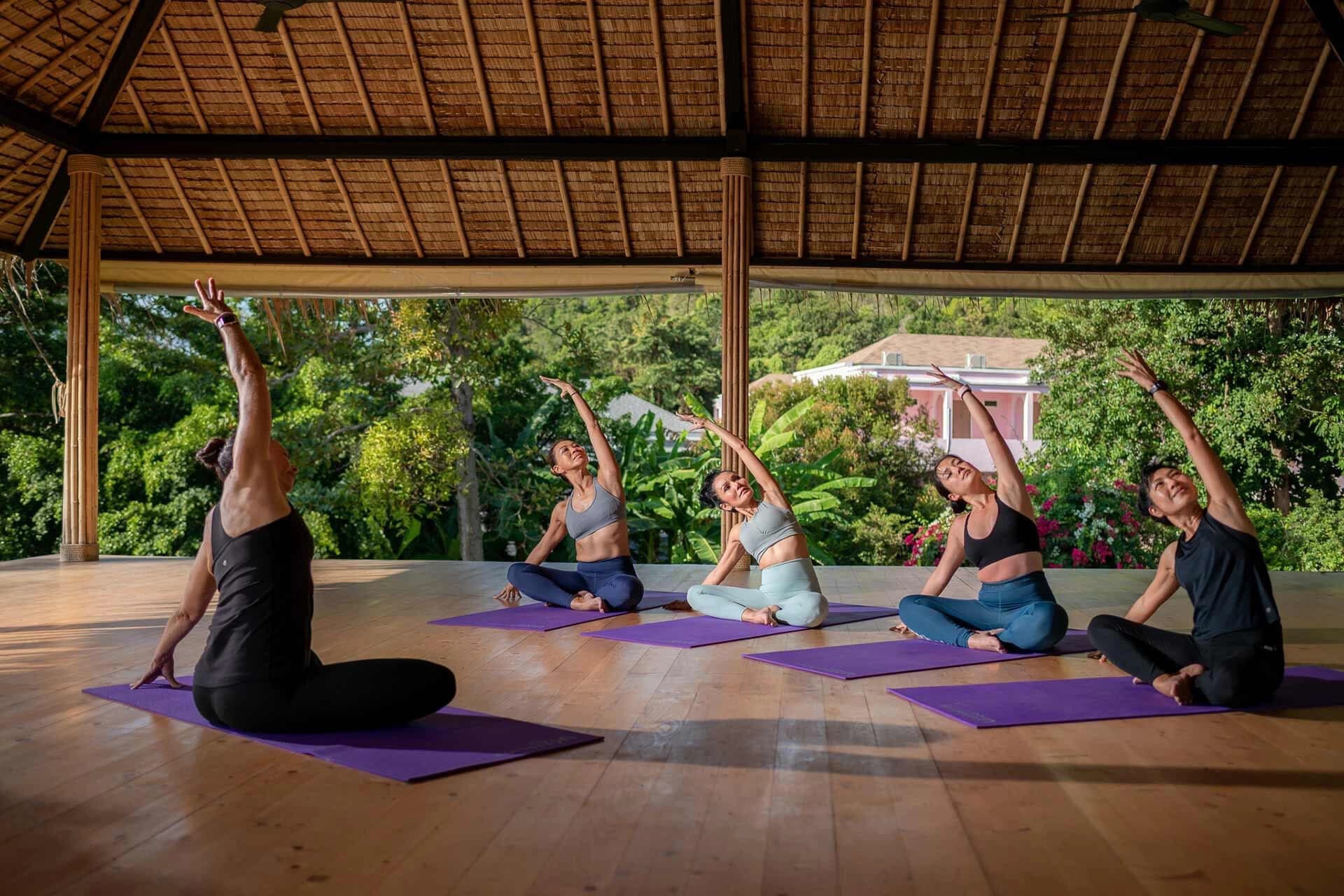 Yoga class with four participants sitting on mats facing yoga instructor with backdrop of Thai plants