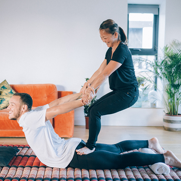 A Thai massage therapist demonstrates a typical back stretch on a male client who is lying face up on a Thai massage mat