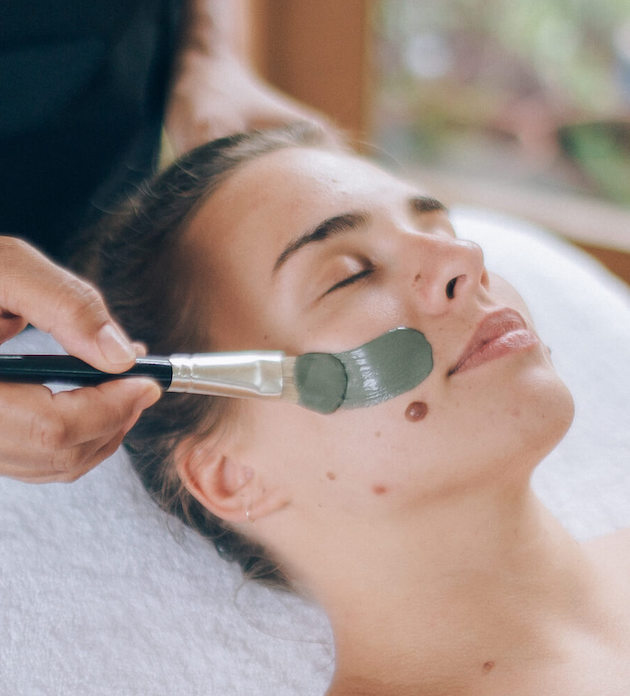Green clay face mask is applied to client's face with a brush as she relaxes on a massage table with her eyes closed